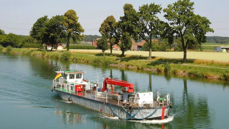 A boat named 'Teuto' on a calm river surrounded by greenery and houses.