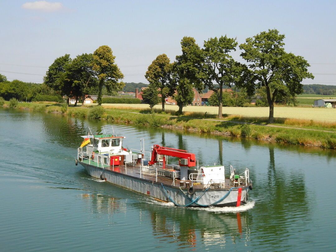 A boat named 'Teuto' on a calm river surrounded by greenery and houses.