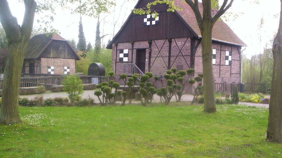 A traditional wooden building with a steeply pitched roof, surrounded by a well-maintained garden with trimmed hedges and flowering plants.