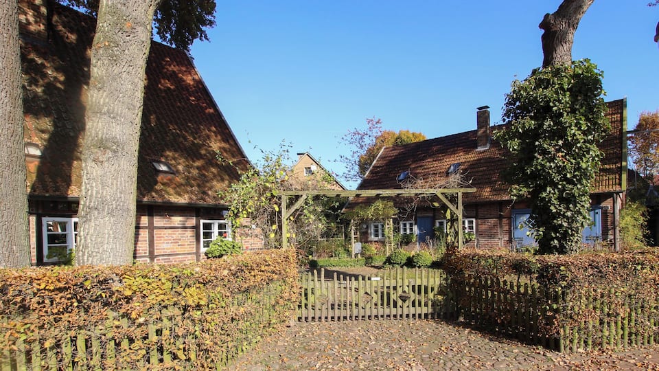 A cobblestone path leading to a wooden gate, surrounded by brick houses with tiled roofs and a large tree in the foreground.