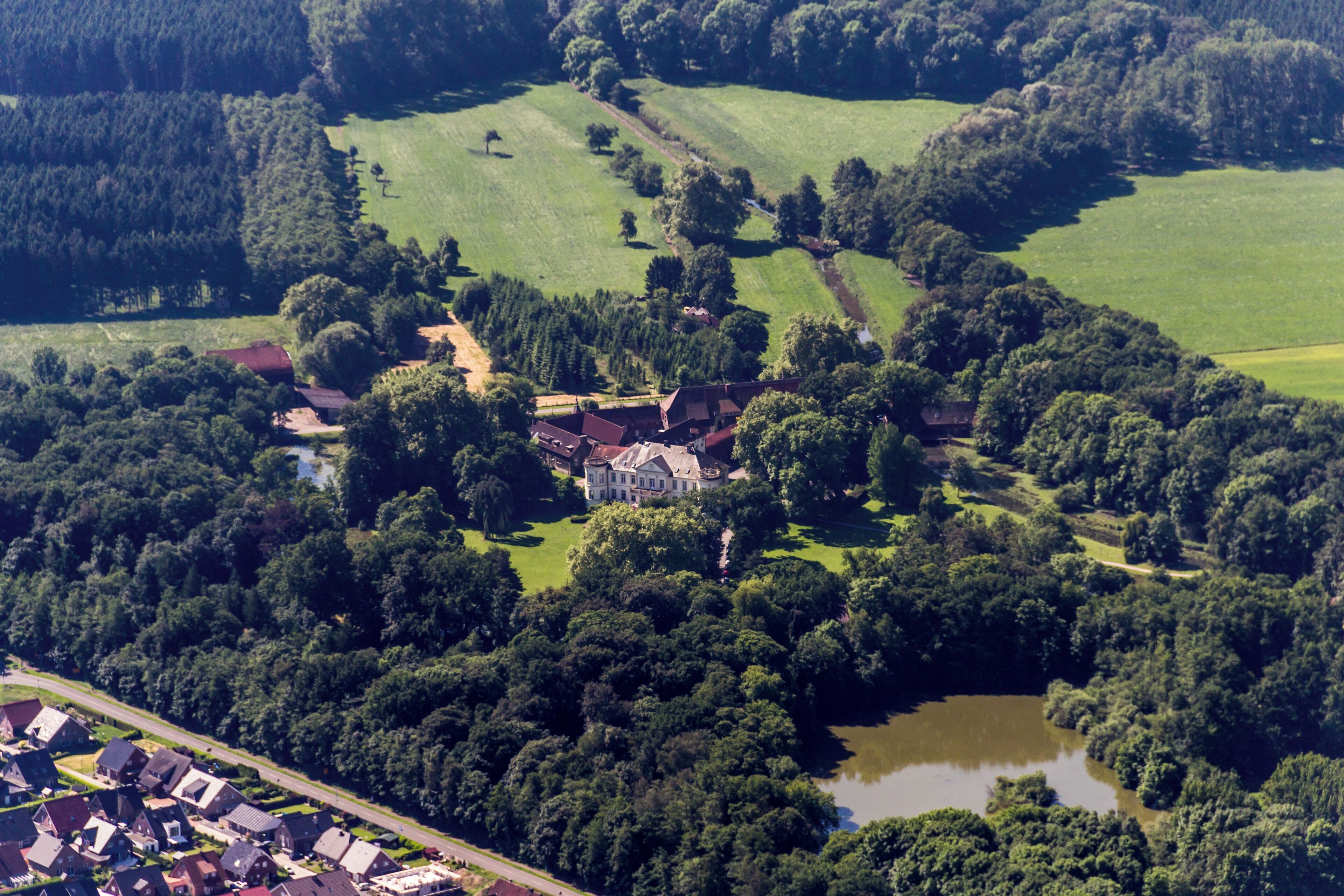 An aerial view of a rural area with a large estate surrounded by dense forests, a small lake, and scattered houses.