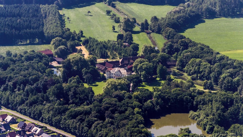An aerial view of a rural area with a large estate surrounded by dense forests, a small lake, and scattered houses.