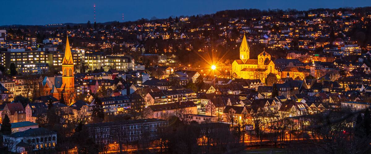 Ein Stadtbild in der Dämmerung mit beleuchteten Gebäuden, einer markanten Kirche mit hohem Turm und einem Glockenturm.