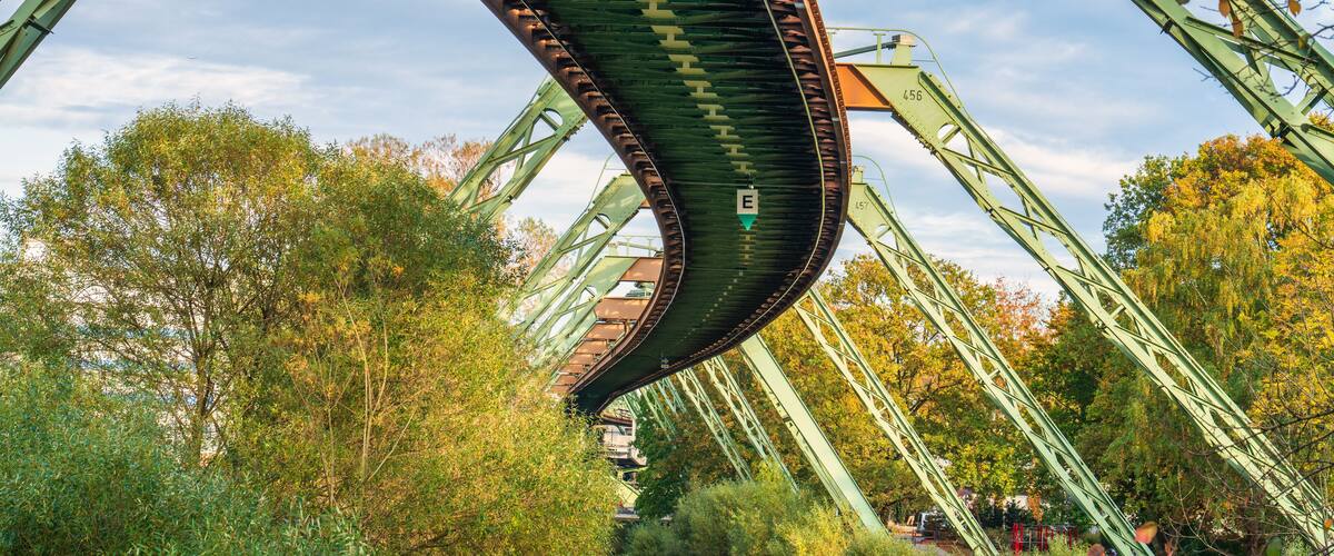Eine grüne Stahlbrücke mit gewölbter Form, umgeben von Bäumen und blauem Himmel.