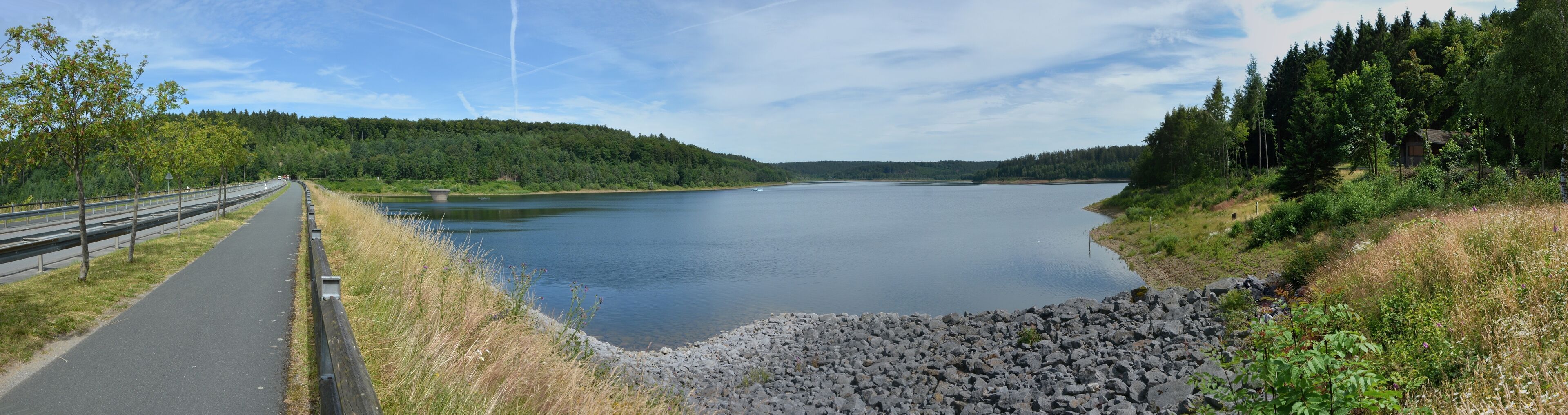 A calm lake with a paved path on the left, surrounded by dense forests and rocky shores.