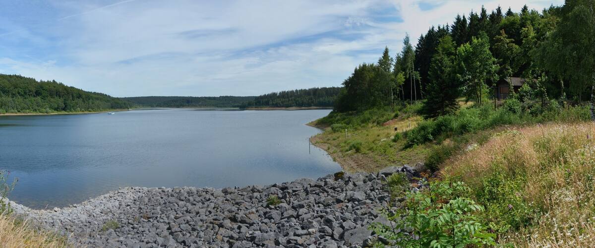 A calm lake with a paved path on the left, surrounded by dense forests and rocky shores.