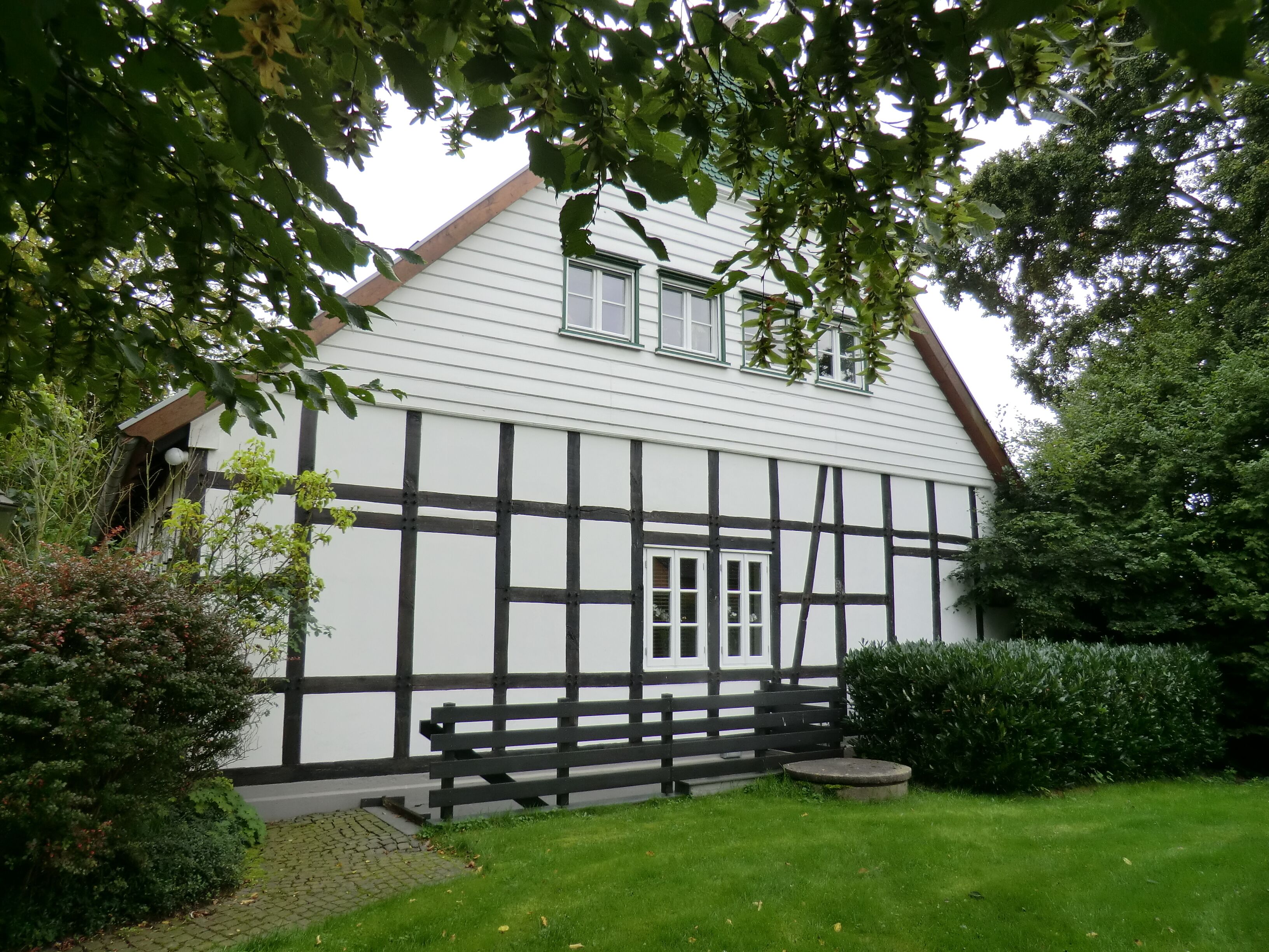 A traditional half-timbered house with a white facade and black timber framing, surrounded by lush greenery and a well-maintained lawn.