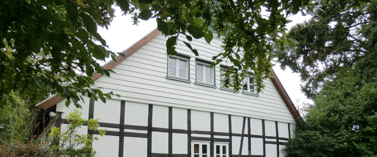 A traditional half-timbered house with a white facade and black timber framing, surrounded by lush greenery and a well-maintained lawn.