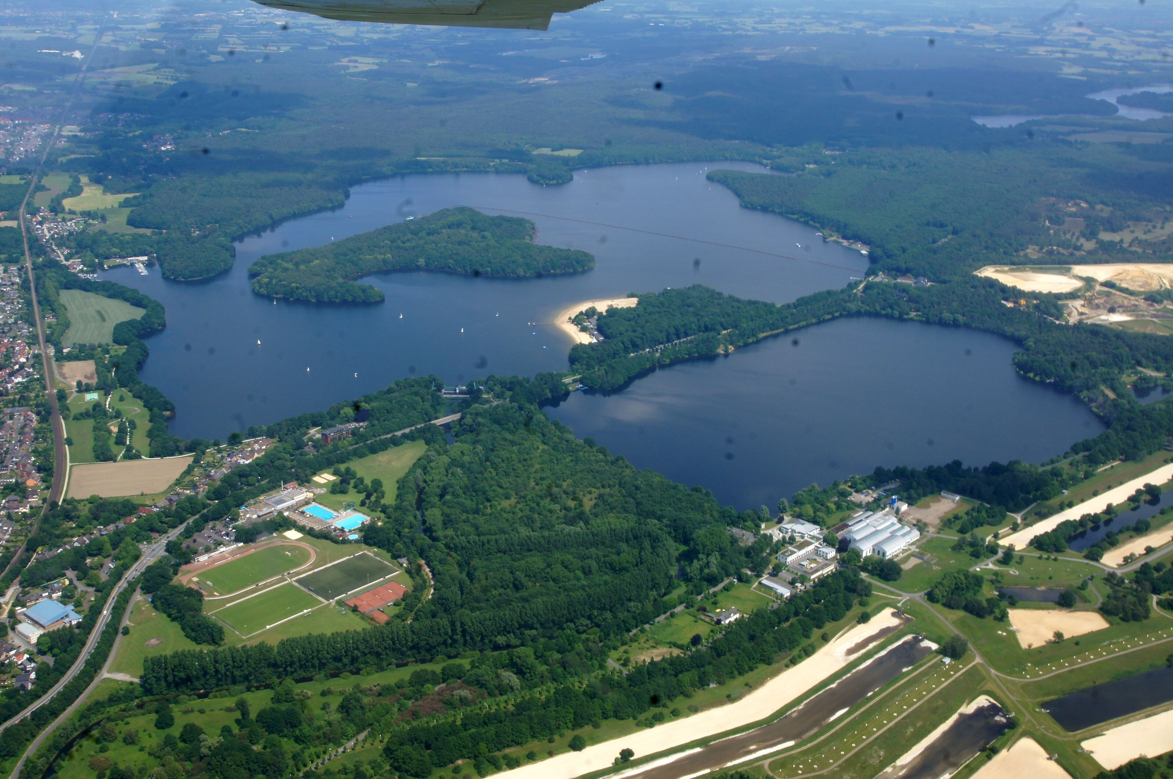 An aerial view of a lake surrounded by forested areas and a small town with visible buildings and roads.