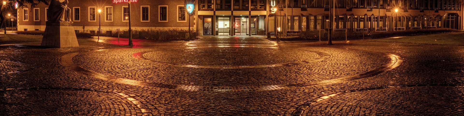 A cobblestone square with a statue, a building with 'RATHAUS' sign, and a 'Sparksse' sign.