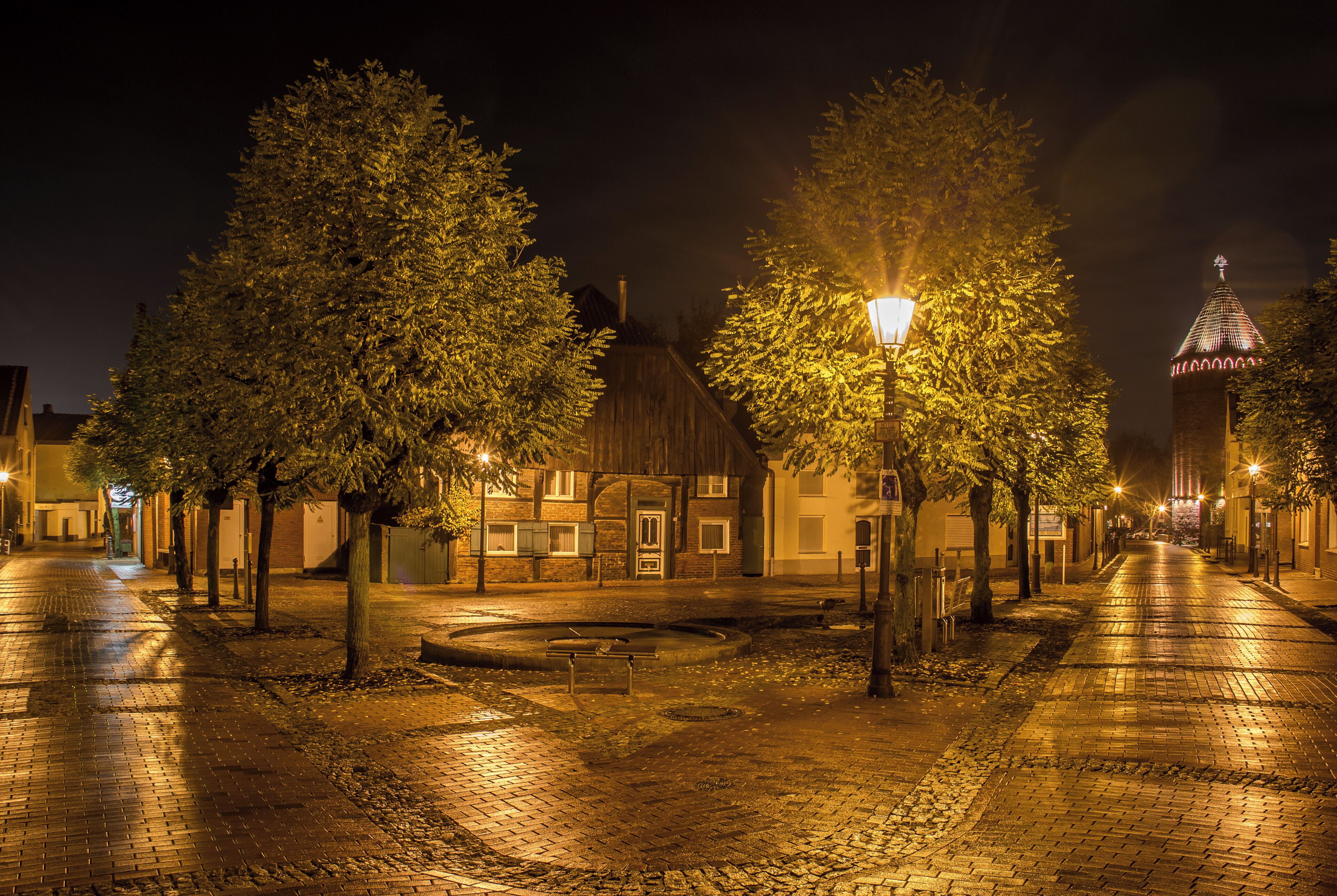 A cobblestone street at night with trees, a bench, and a building with a pointed roof.