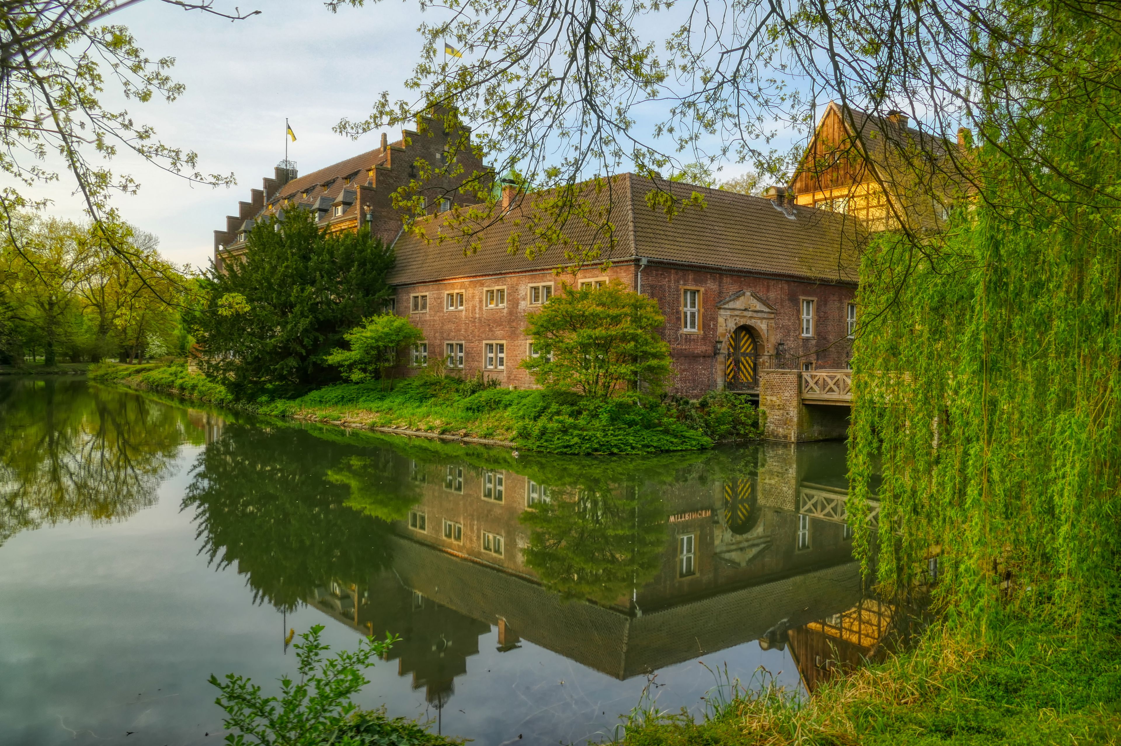 A historic building with a reflection in the water, surrounded by lush greenery and trees.