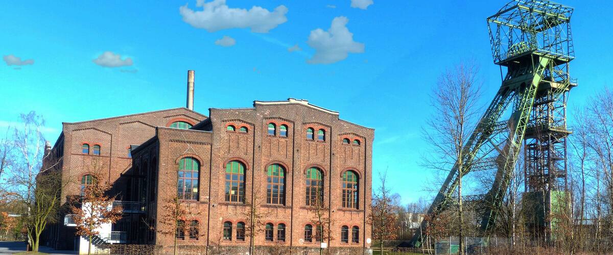 A large brick building with arched windows and a tall chimney, adjacent to a large industrial structure with a crane.