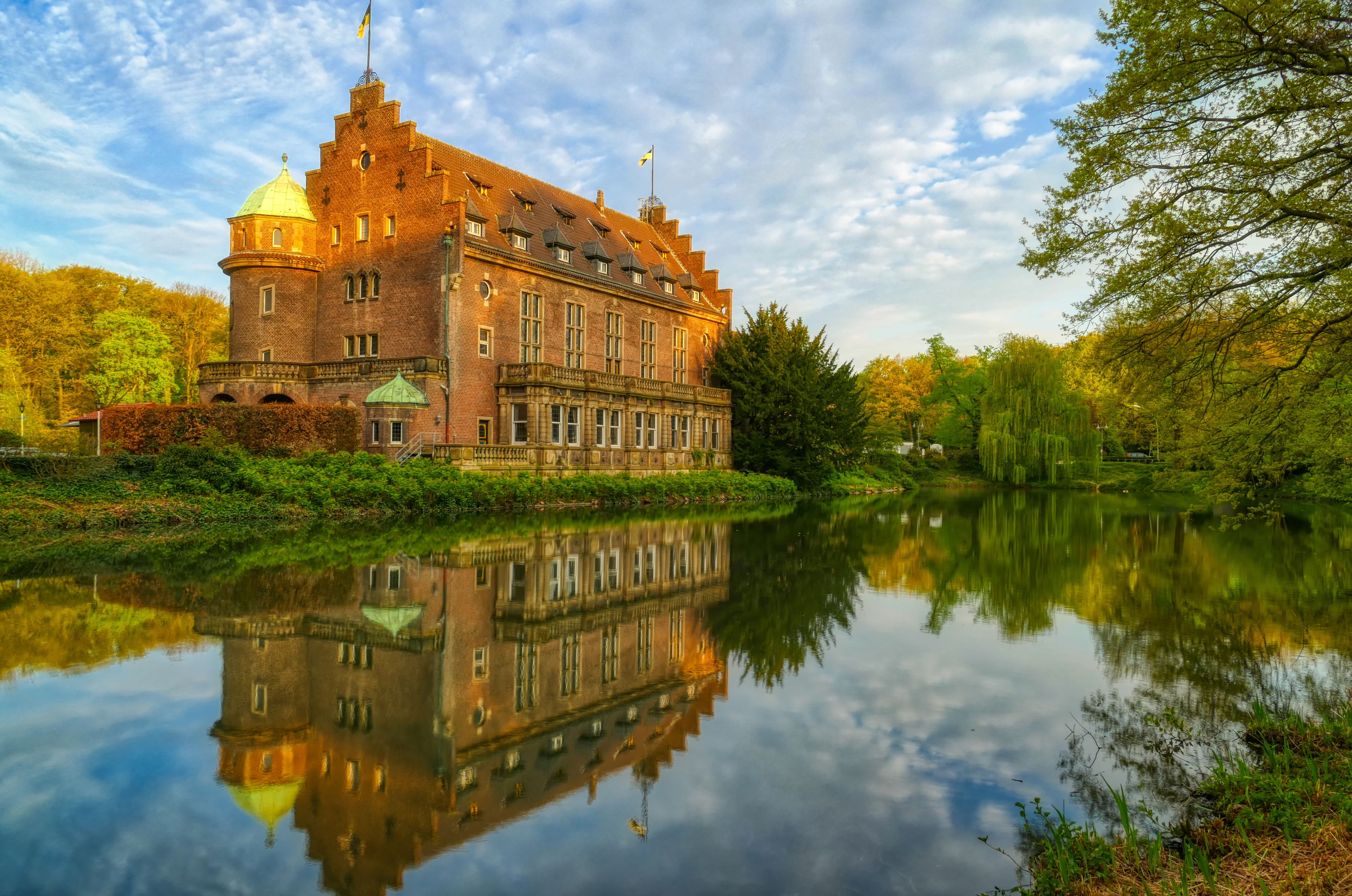 A large, historic brick building with a reflection in a calm body of water, surrounded by lush greenery and trees.