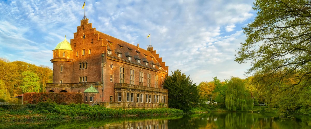 A large, historic brick building with a reflection in a calm body of water, surrounded by lush greenery and trees.