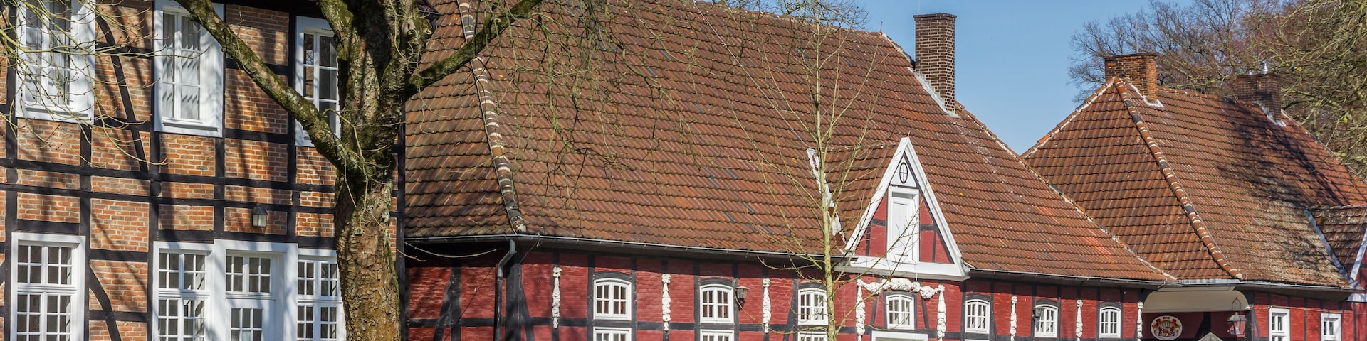 Ein traditionelles Fachwerkhäuschen mit roter Fassade und weißen Fensterrahmen, umgeben von einem gepflegten Rasen und alten Bäumen.