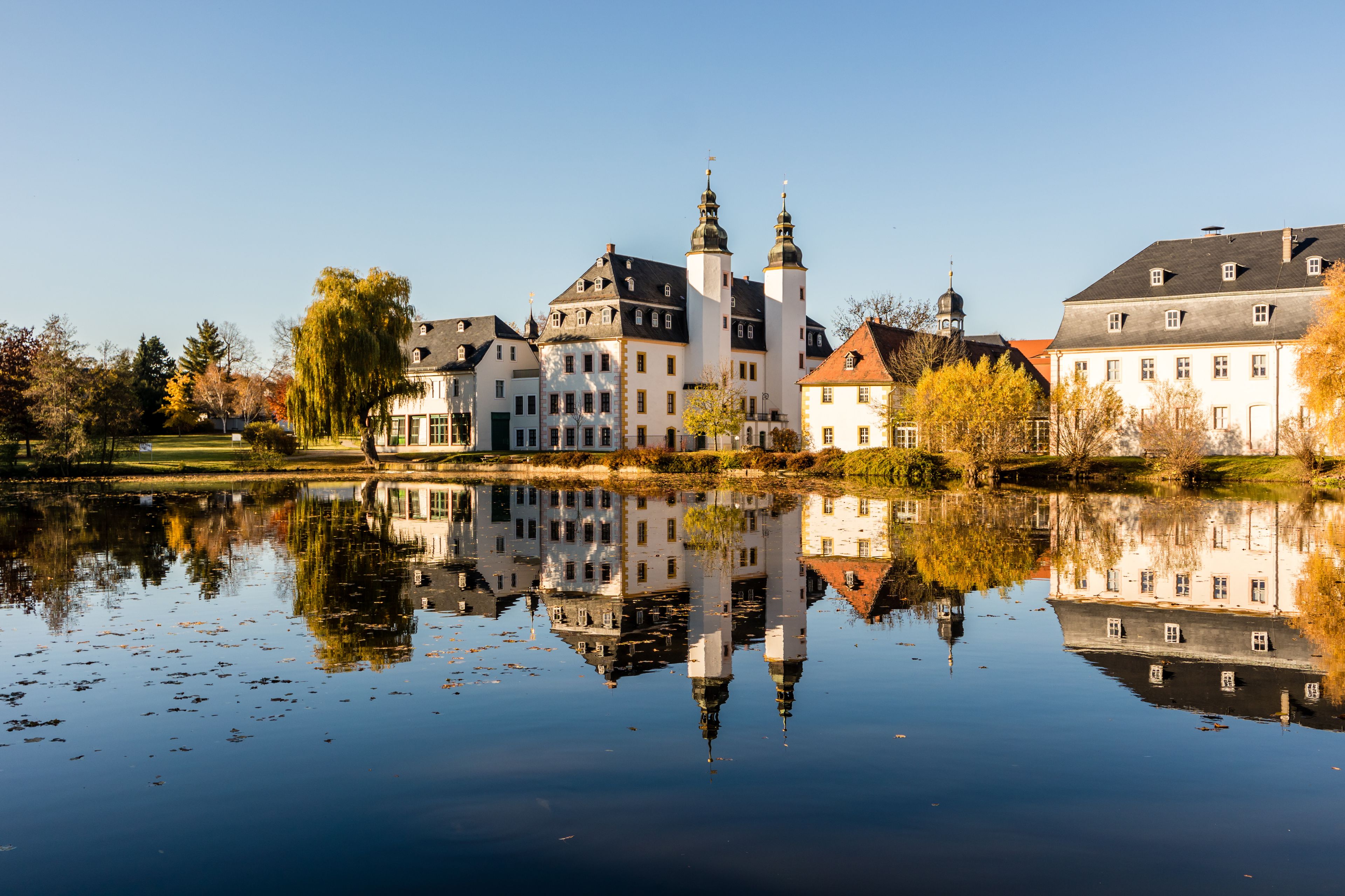 A historic castle with multiple towers, a calm lake reflecting the buildings, and autumn trees in the foreground.