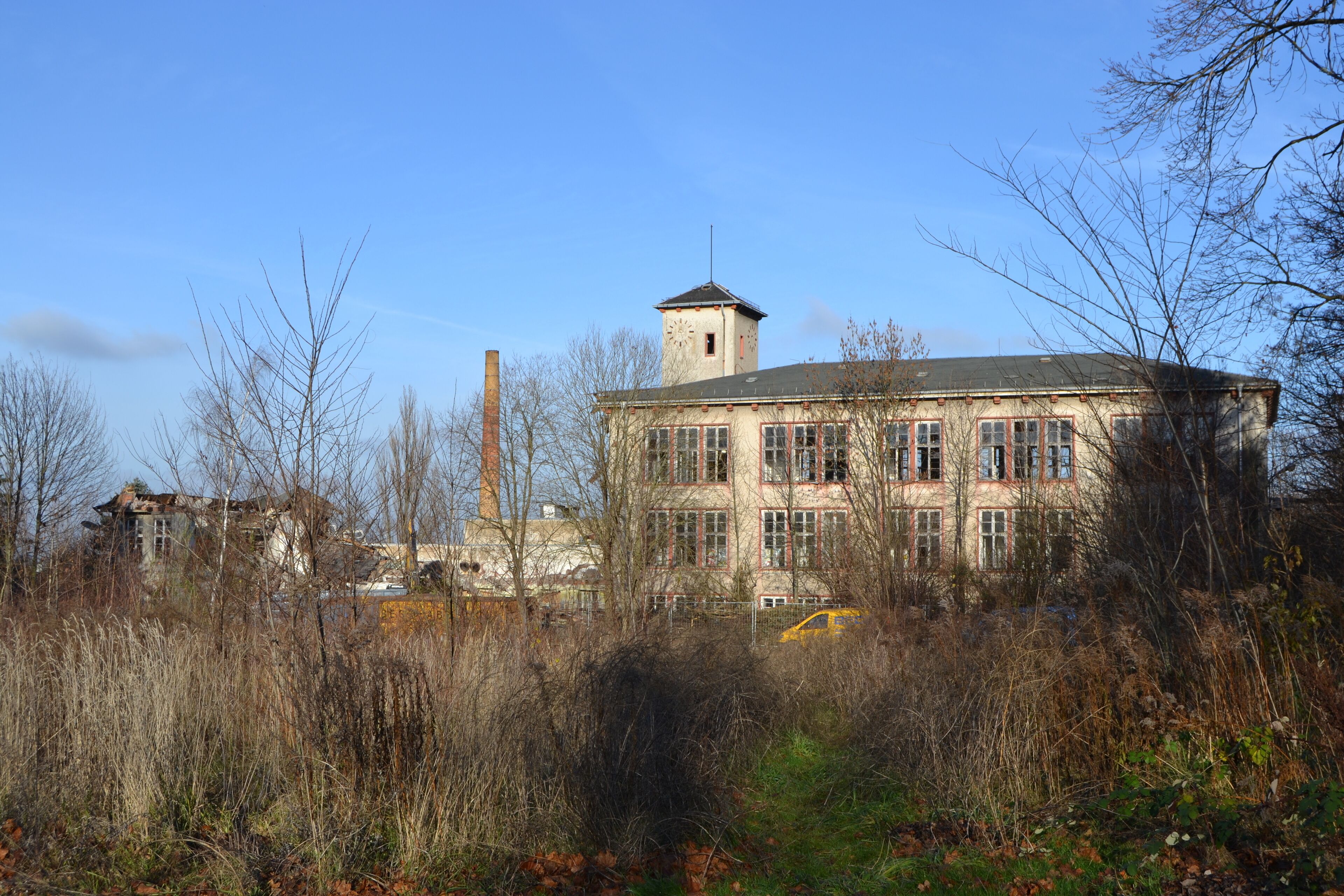 An abandoned building with a clock tower, surrounded by overgrown vegetation and a yellow car in the foreground.