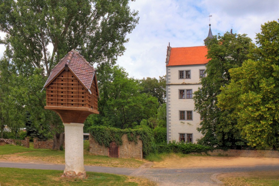 A birdhouse on a white pedestal, a building with a red roof, and lush green trees.