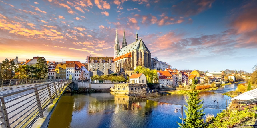 Un río con un puente, una iglesia histórica con dos torres gemelas y edificios circundantes.