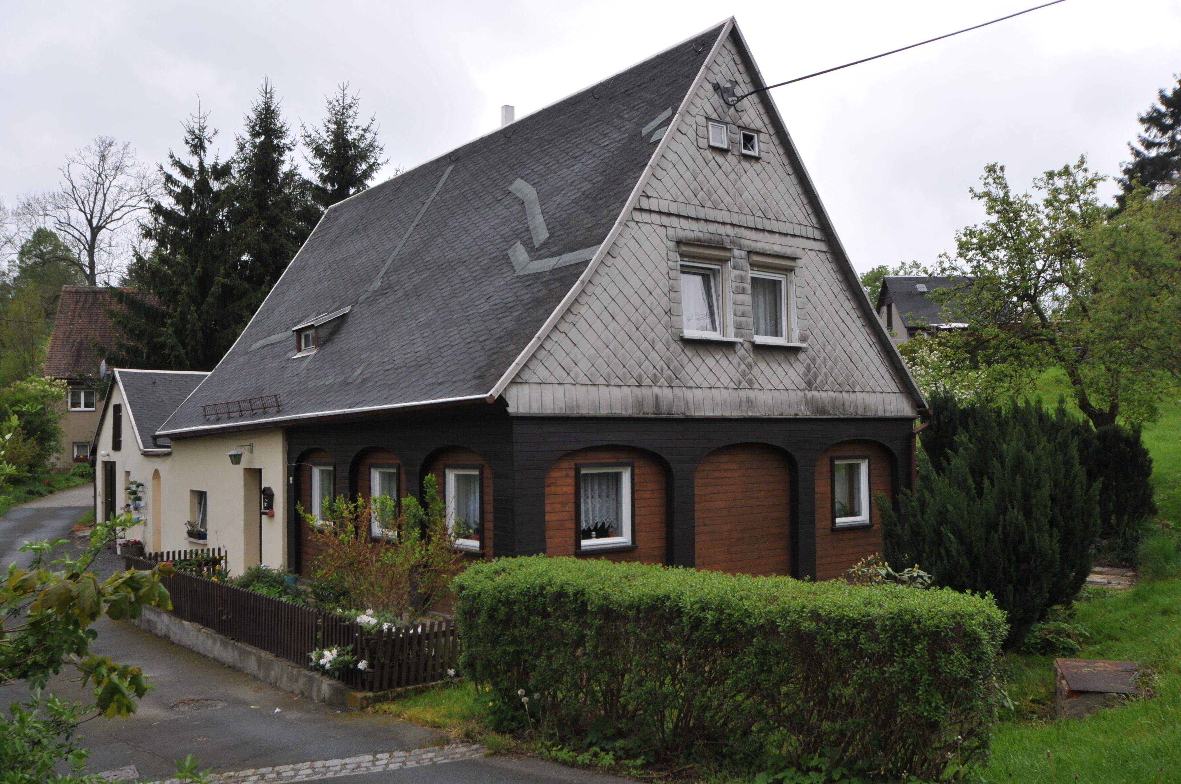 A house with a steeply pitched roof, a small attached garage, and a well-maintained hedge in front.
