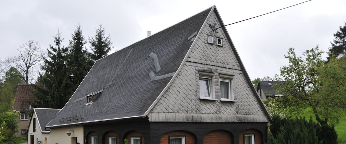 A house with a steeply pitched roof, a small attached garage, and a well-maintained hedge in front.