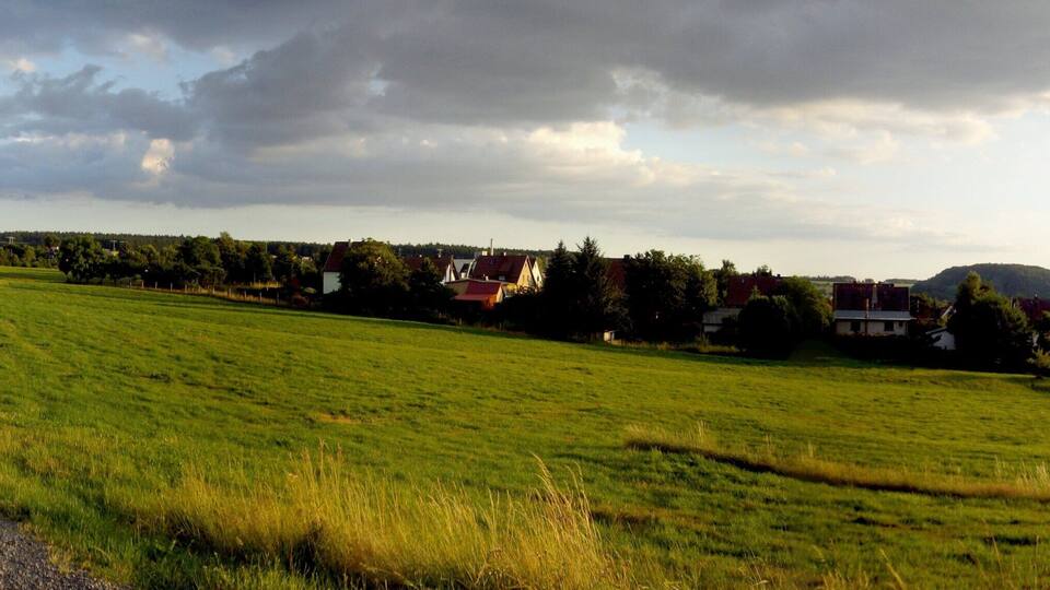 A rural landscape with a gravel path, green fields, houses, and a hill in the distance.