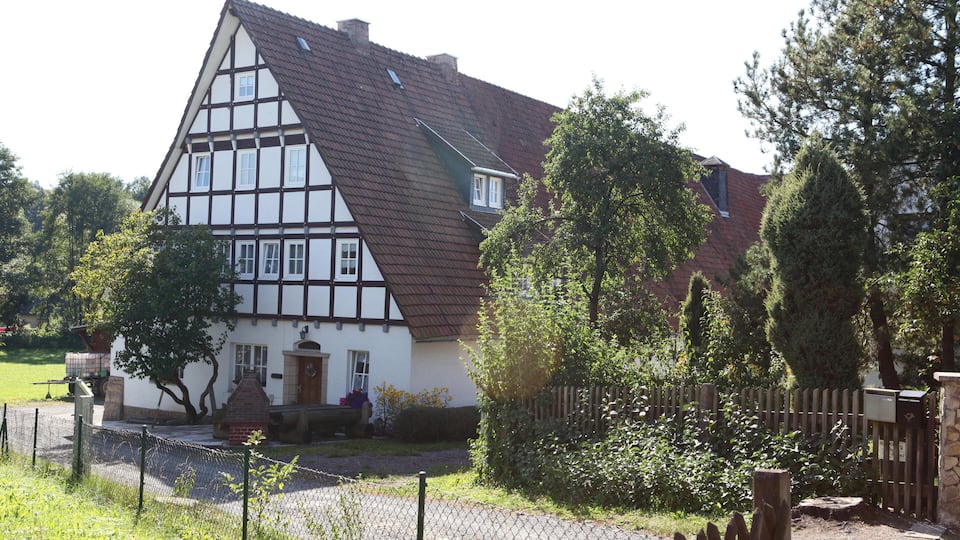 A traditional half-timbered house with a sloped roof, surrounded by trees and a wooden fence.