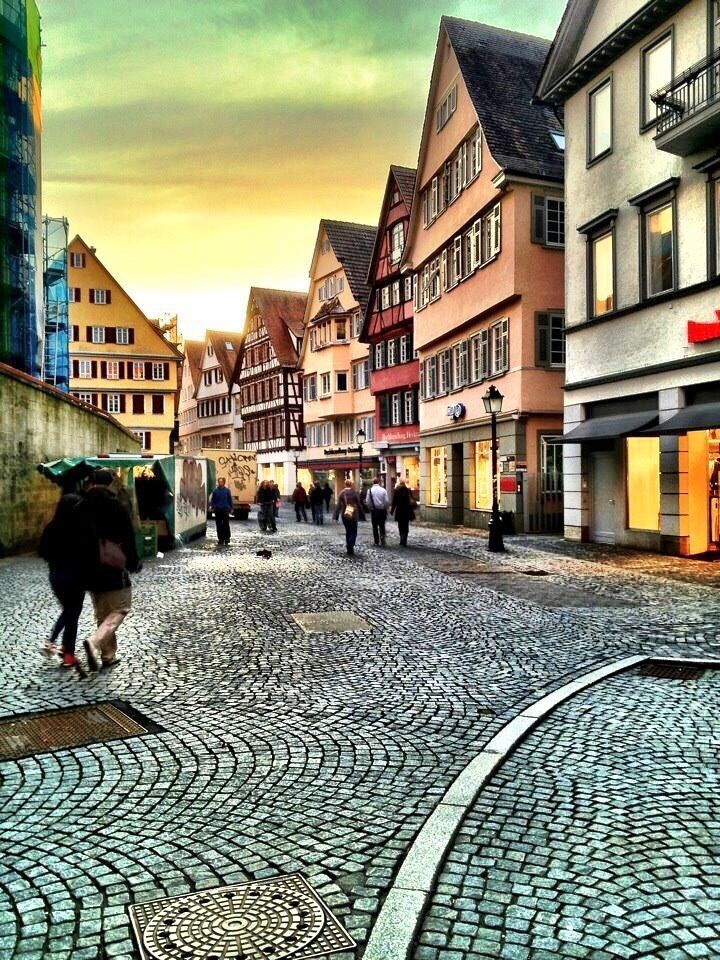 A cobblestone street lined with traditional half-timbered buildings, pedestrians, and a modern building with scaffolding.