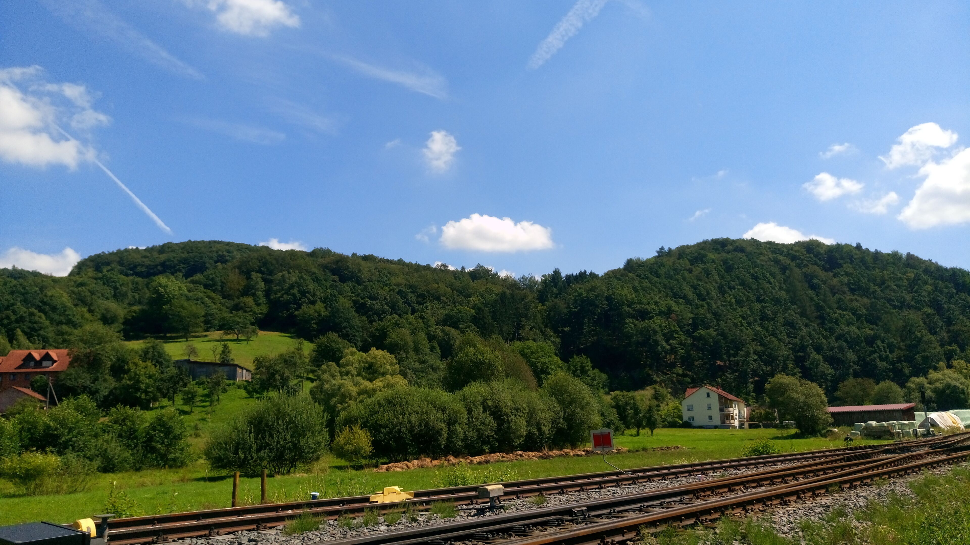 A rural landscape with a railway track, green fields, and a hill with trees.