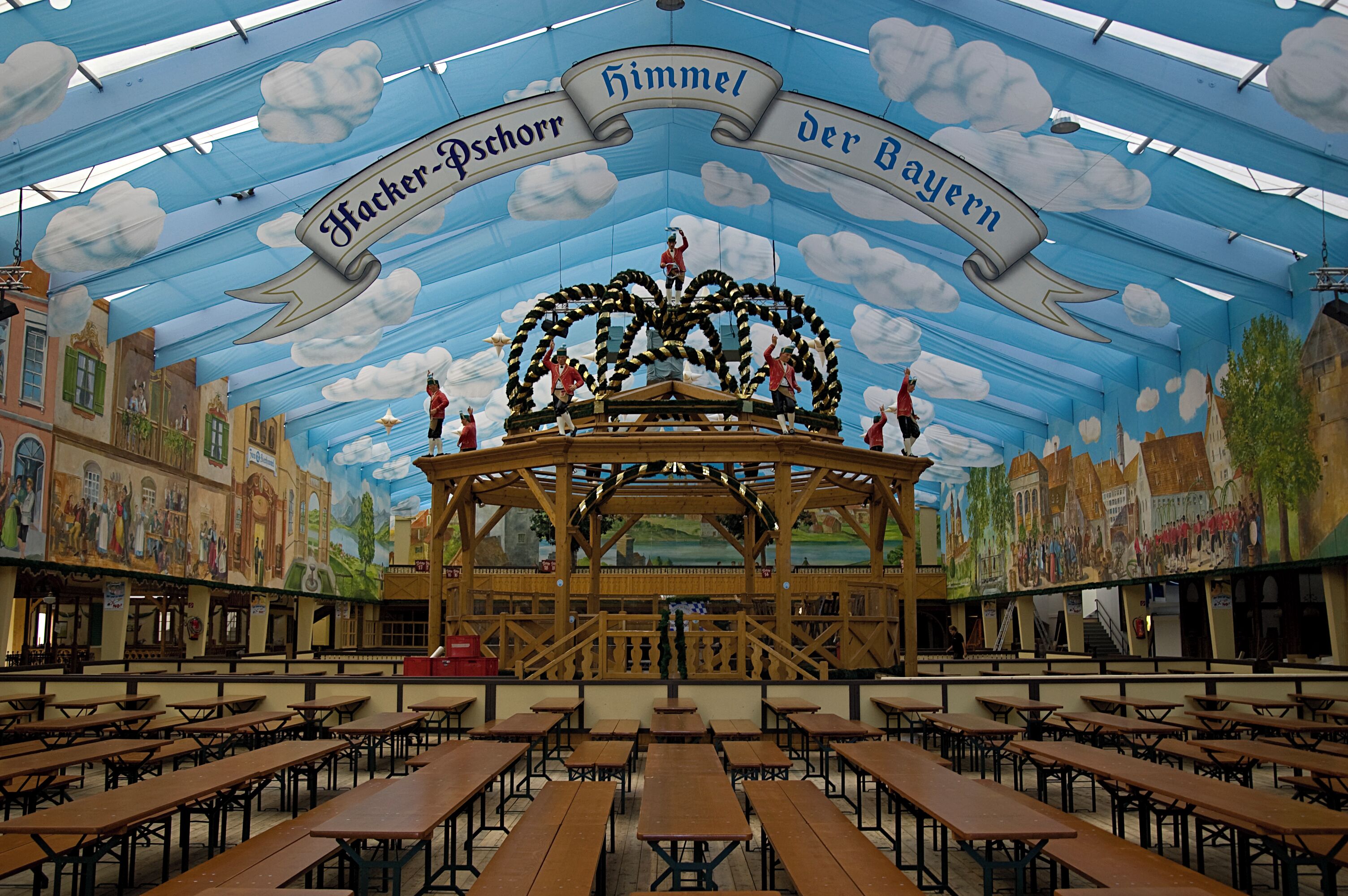 A large hall with wooden tables and a stage with a banner reading "Hacker-Pschorr der Bayern".