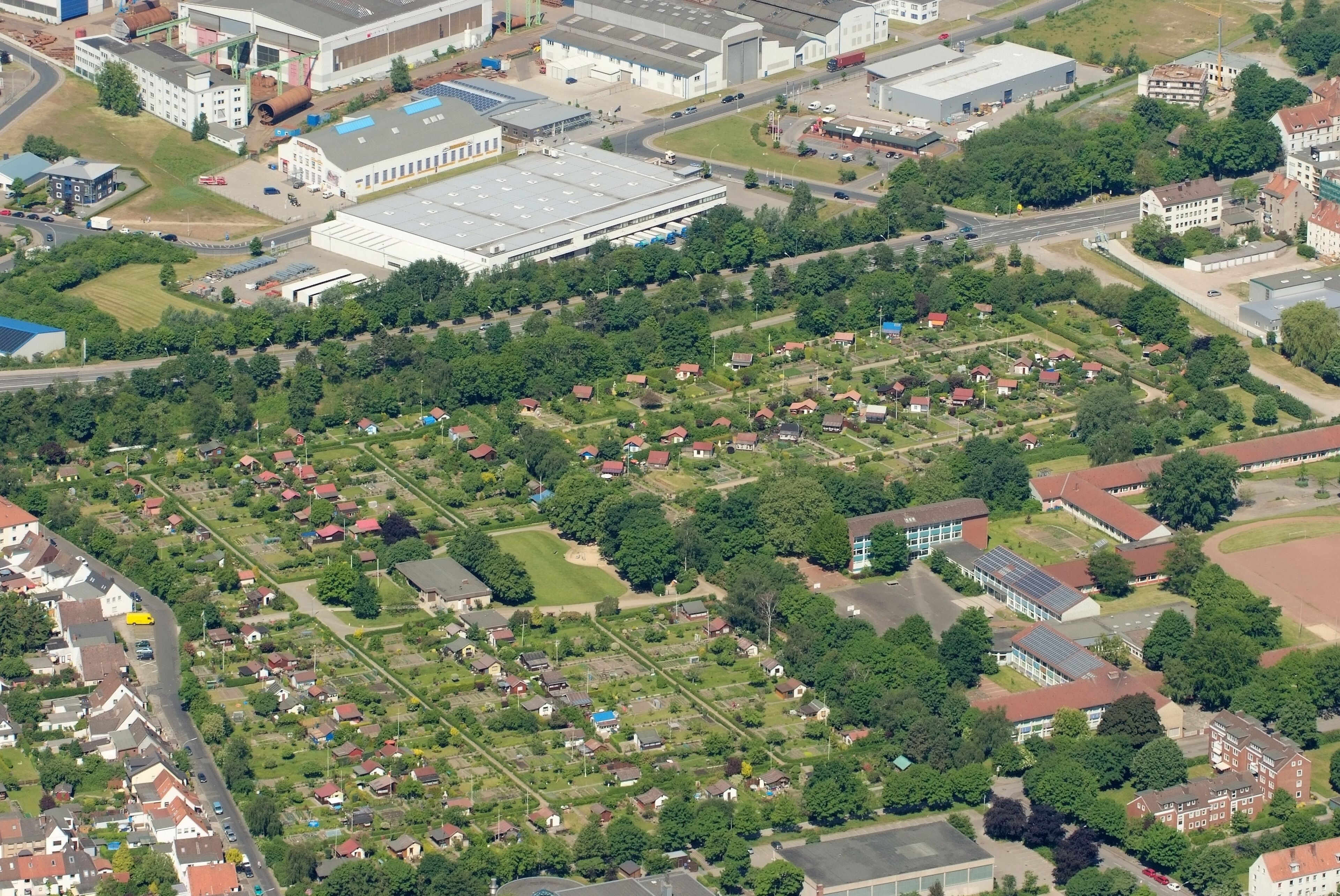 An aerial view of a residential area with houses, a large industrial building, and surrounding greenery.