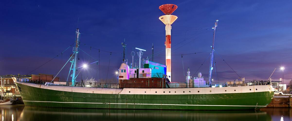 A large green ship docked at a harbor with a tall, illuminated tower in the background.