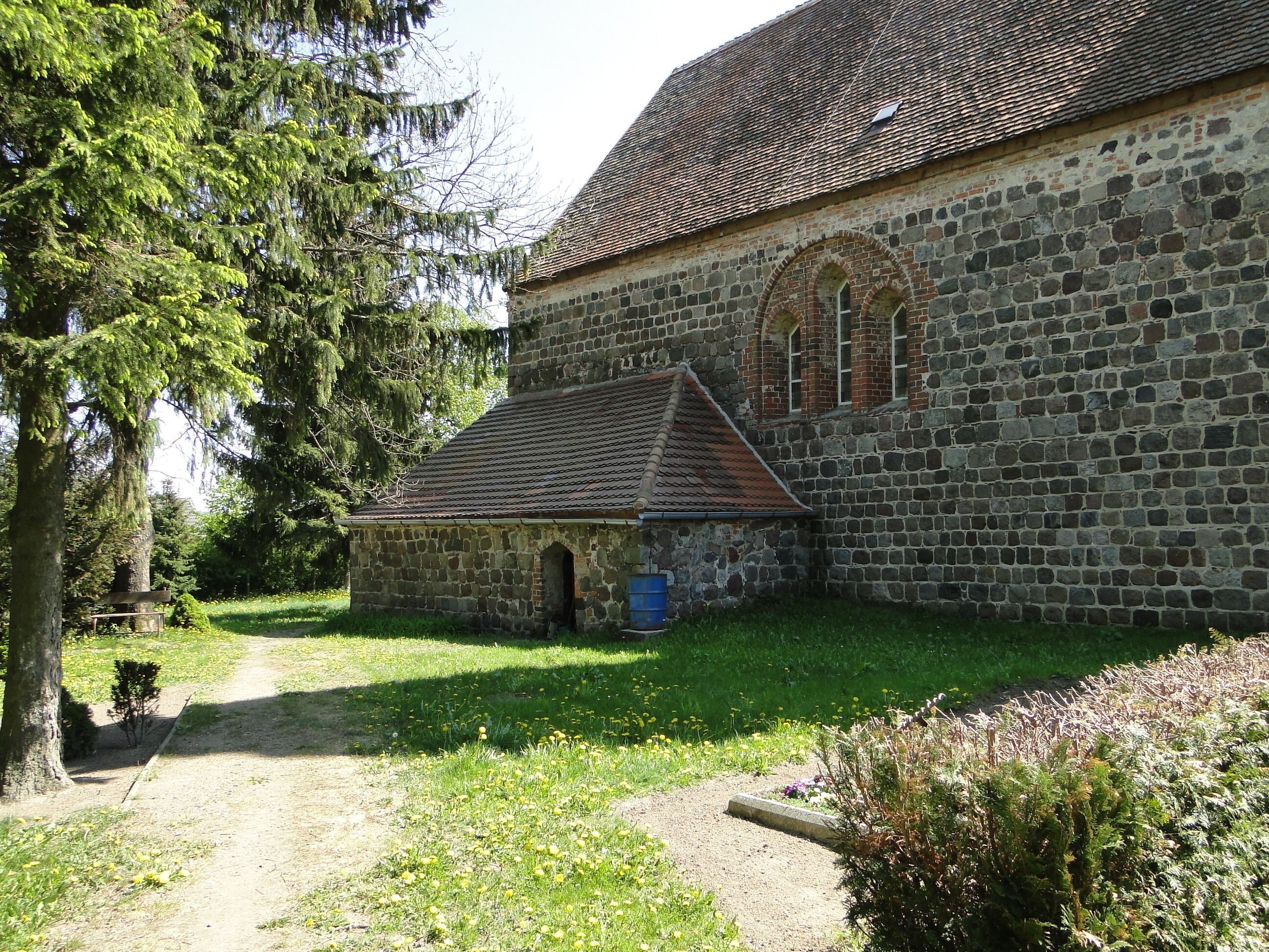 A stone church with a tiled roof and arched windows, surrounded by greenery and a gravel path.
