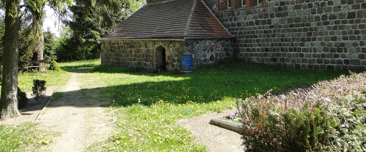 A stone church with a tiled roof and arched windows, surrounded by greenery and a gravel path.