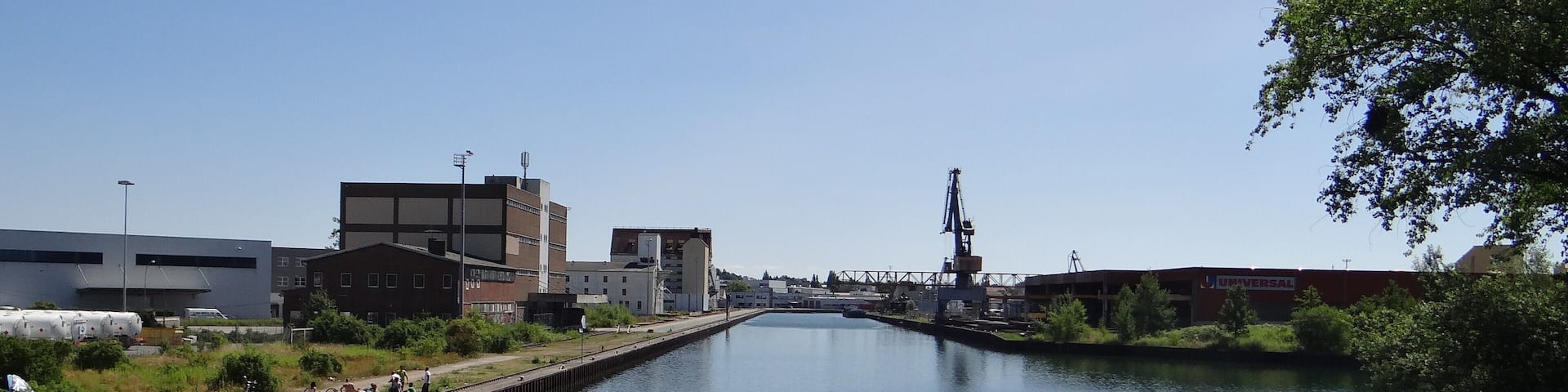 A calm canal with industrial buildings and a bridge in the background.
