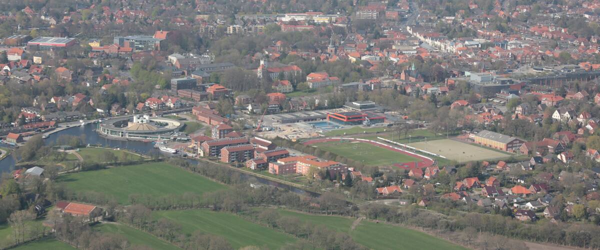 An aerial view of a town with a river, green fields, and red-roofed buildings.