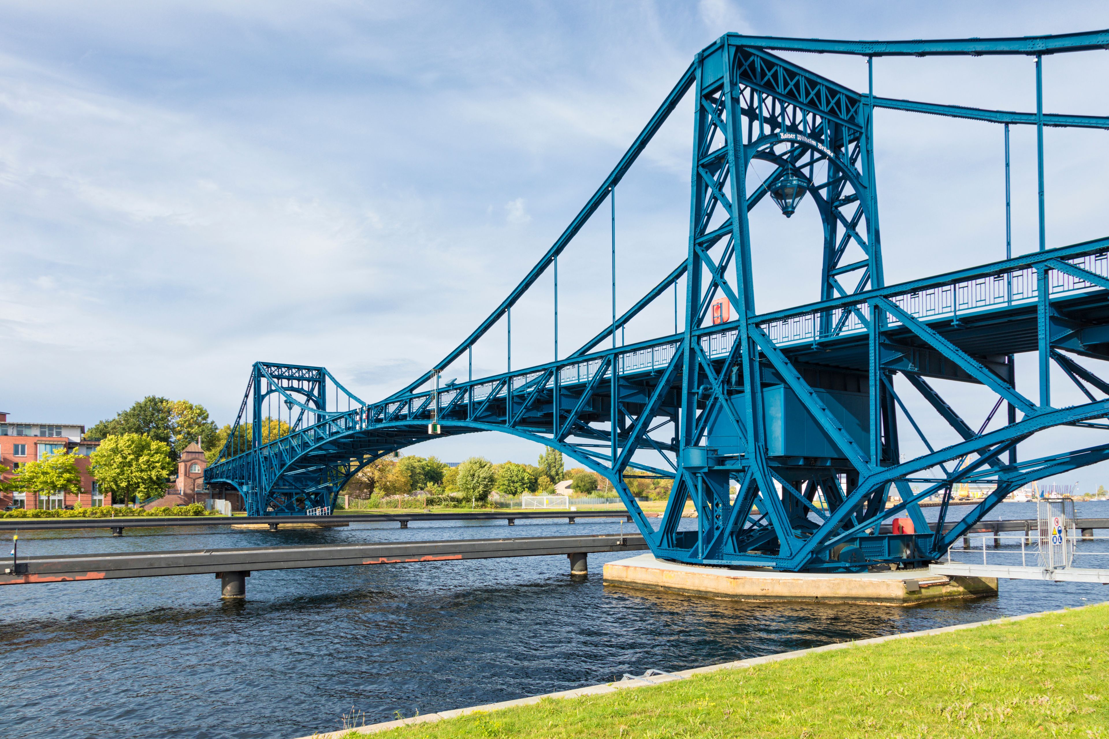 A blue steel bridge over a river with a cityscape in the background.