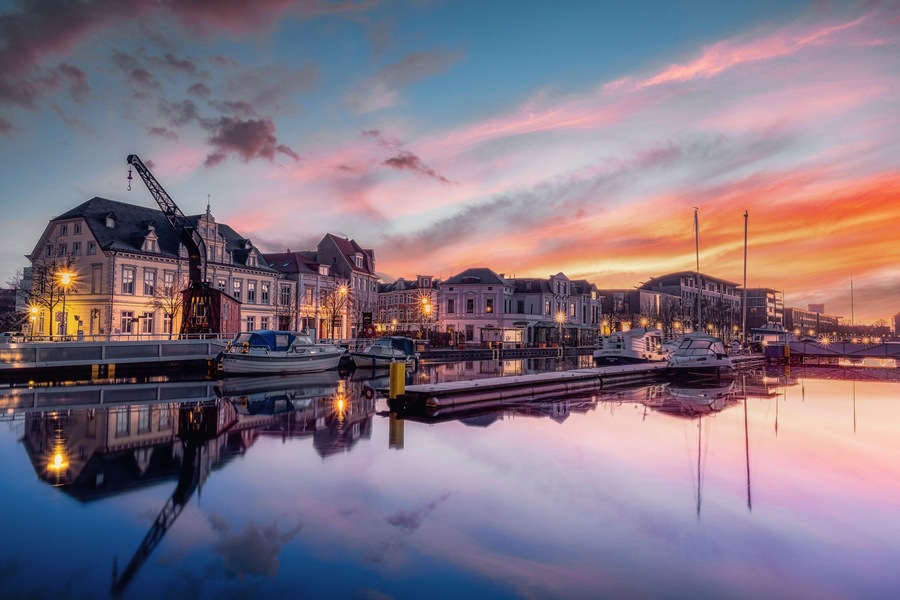 Un paisaje urbano frente al agua al atardecer, con edificios iluminados y barcos amarrados a lo largo del agua tranquila.