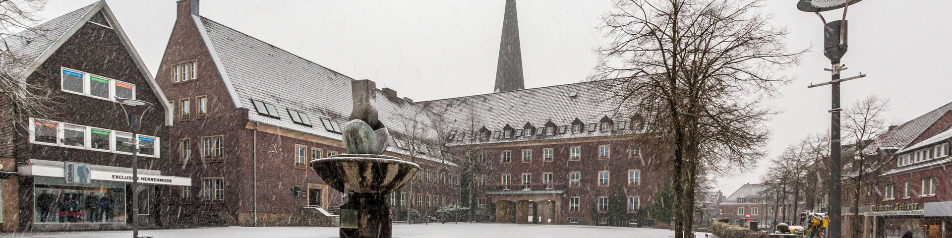 A snow-covered square with a central fountain, brick buildings, and a church spire in the background.
