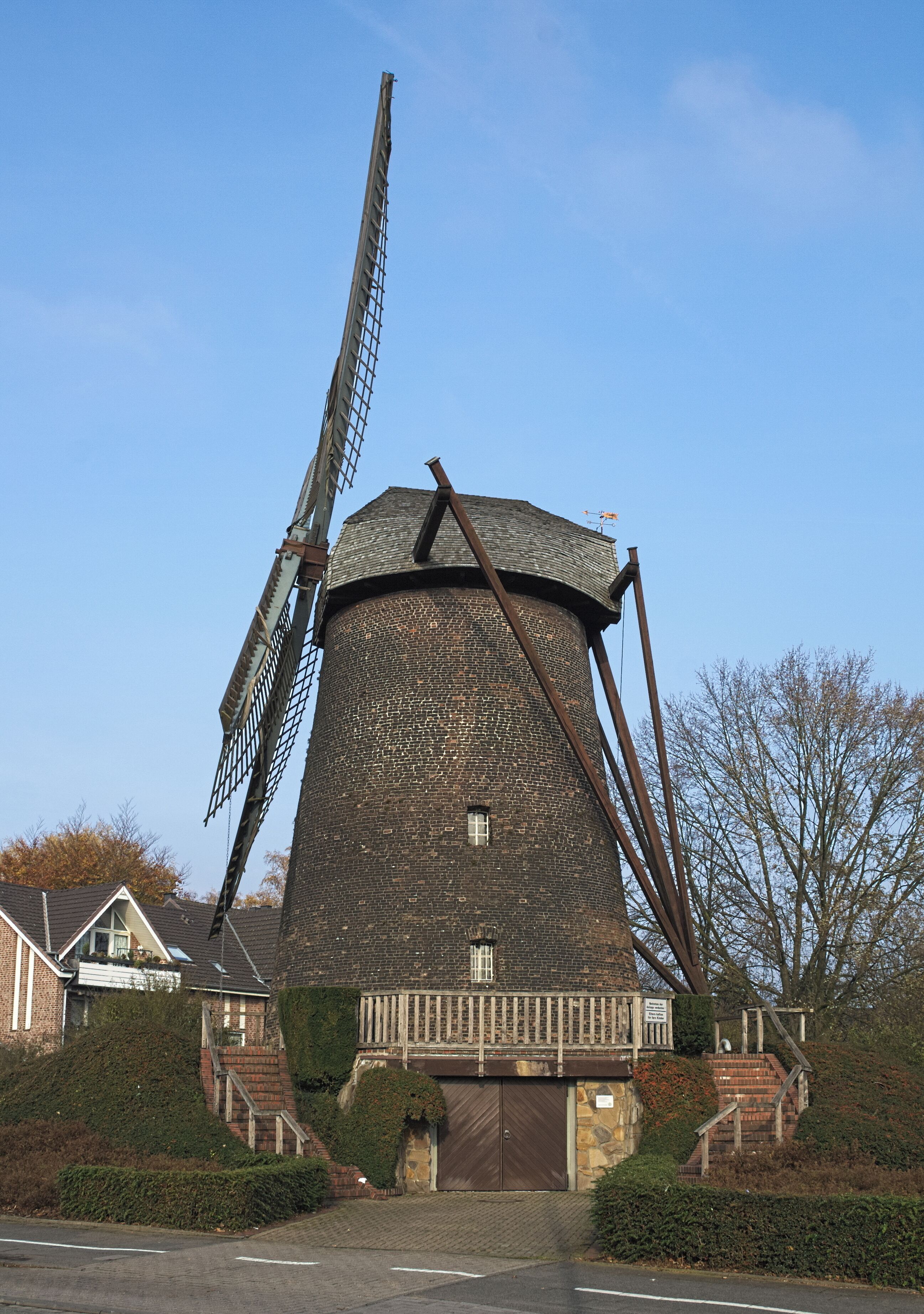 A large windmill with a brick base and a metal structure, surrounded by a residential area with houses and a clear sky.
