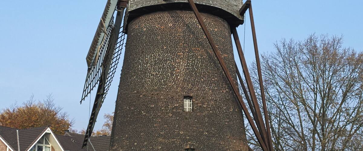 A large windmill with a brick base and a metal structure, surrounded by a residential area with houses and a clear sky.