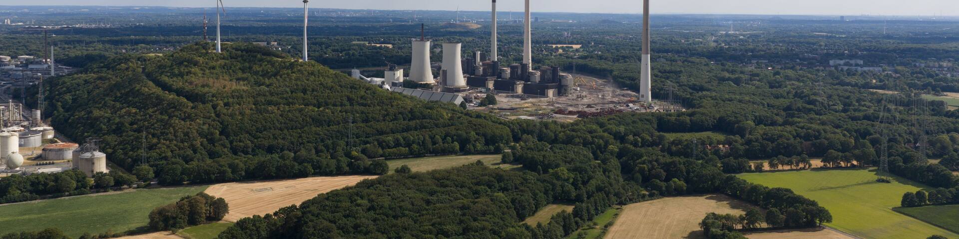 A wind farm with multiple wind turbines and cooling towers amidst a hilly landscape with fields and forests.