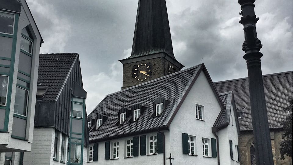 Een kerk met een hoge spits en een klok, omringd door gebouwen met witte gevels en groene luiken.