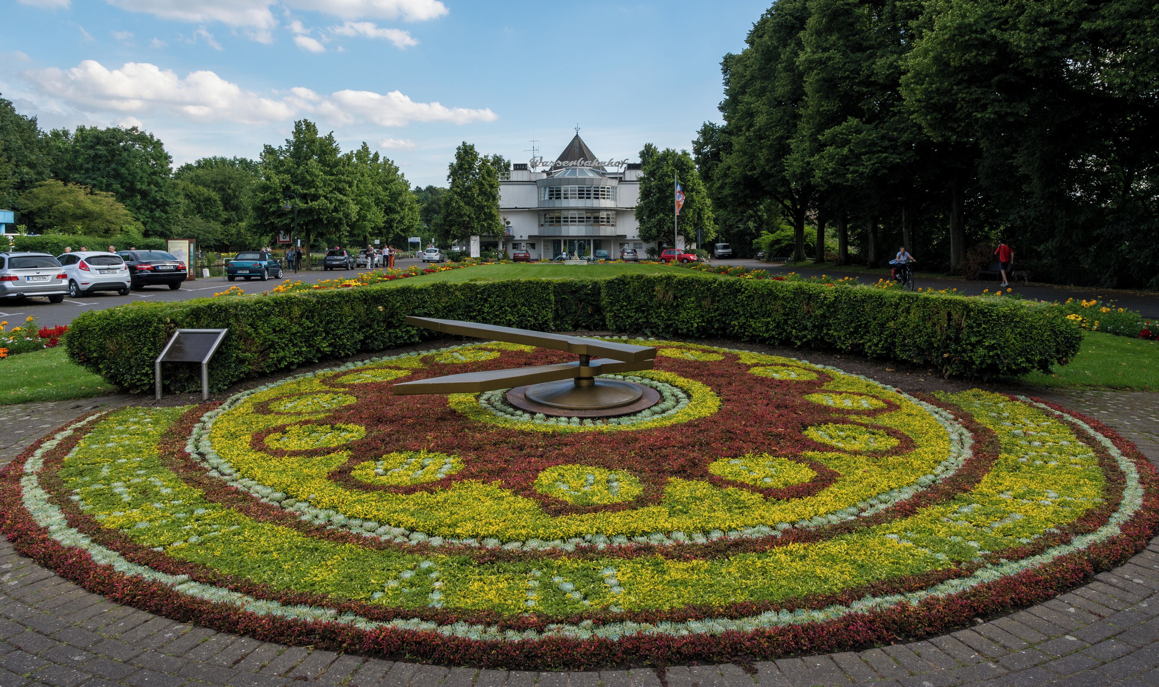 A circular flower bed with a central sculpture, surrounded by a building with a pointed roof and a flag.