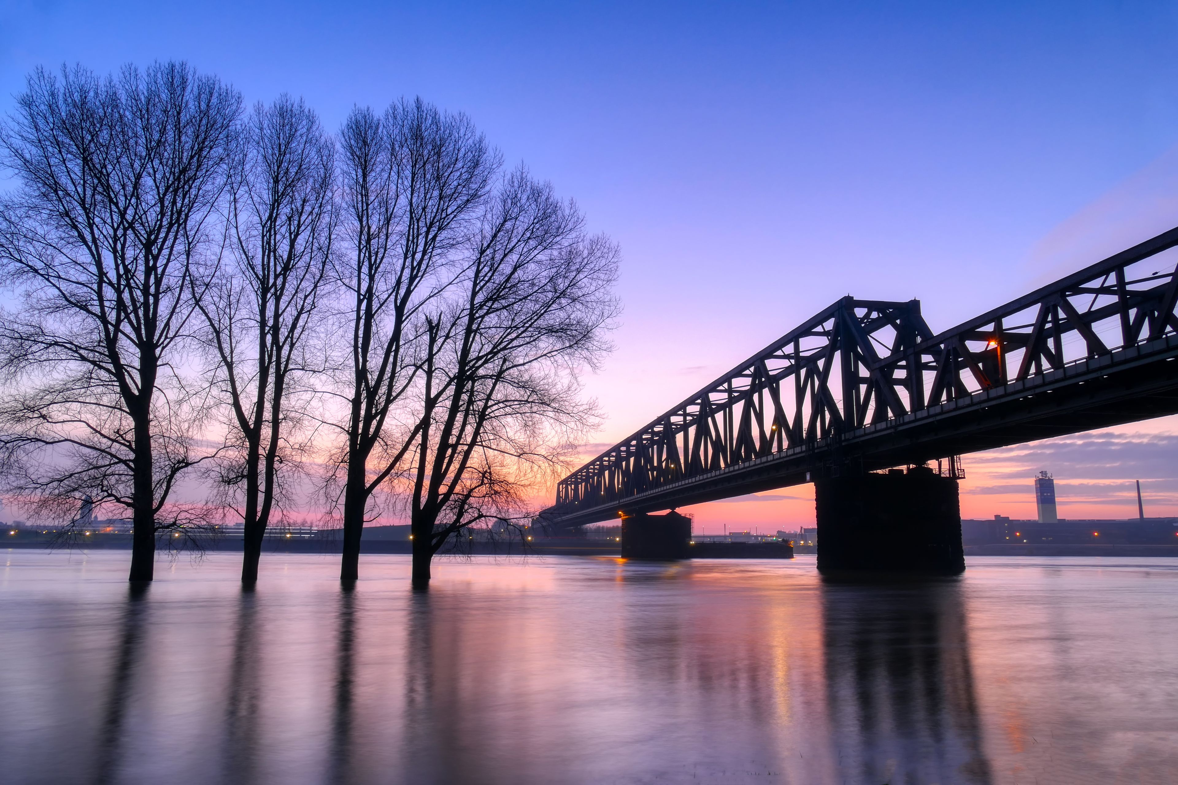 Un puente sobre un río tranquilo al atardecer, con árboles desnudos a la izquierda y el horizonte de una ciudad al fondo.