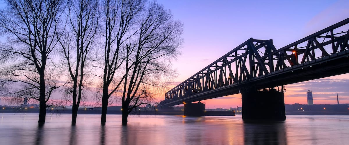 Eine Brücke über einem ruhigen Fluss im Abenddämmerung, mit kahlen Bäumen links und einer Stadtansicht im Hintergrund.