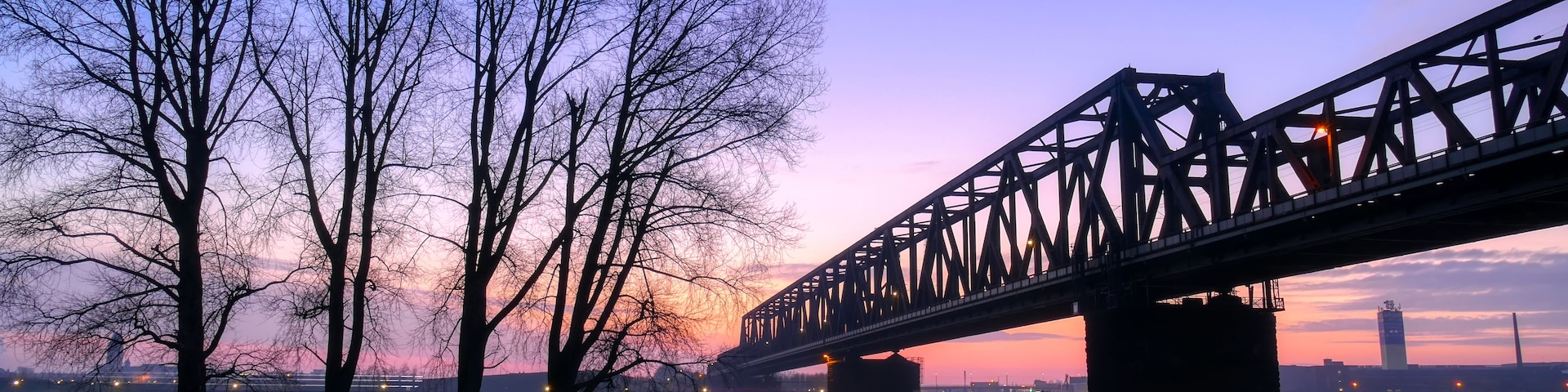 Un puente sobre un río tranquilo al atardecer, con árboles desnudos a la izquierda y el horizonte de una ciudad al fondo.