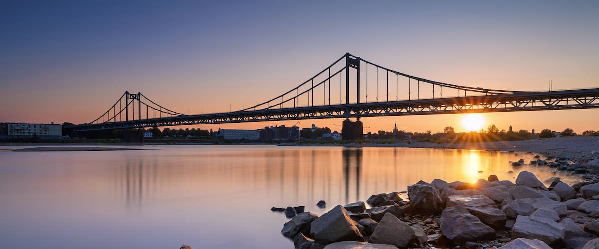 Eine Brücke über einem ruhigen Fluss bei Sonnenuntergang, im Vordergrund Felsen.