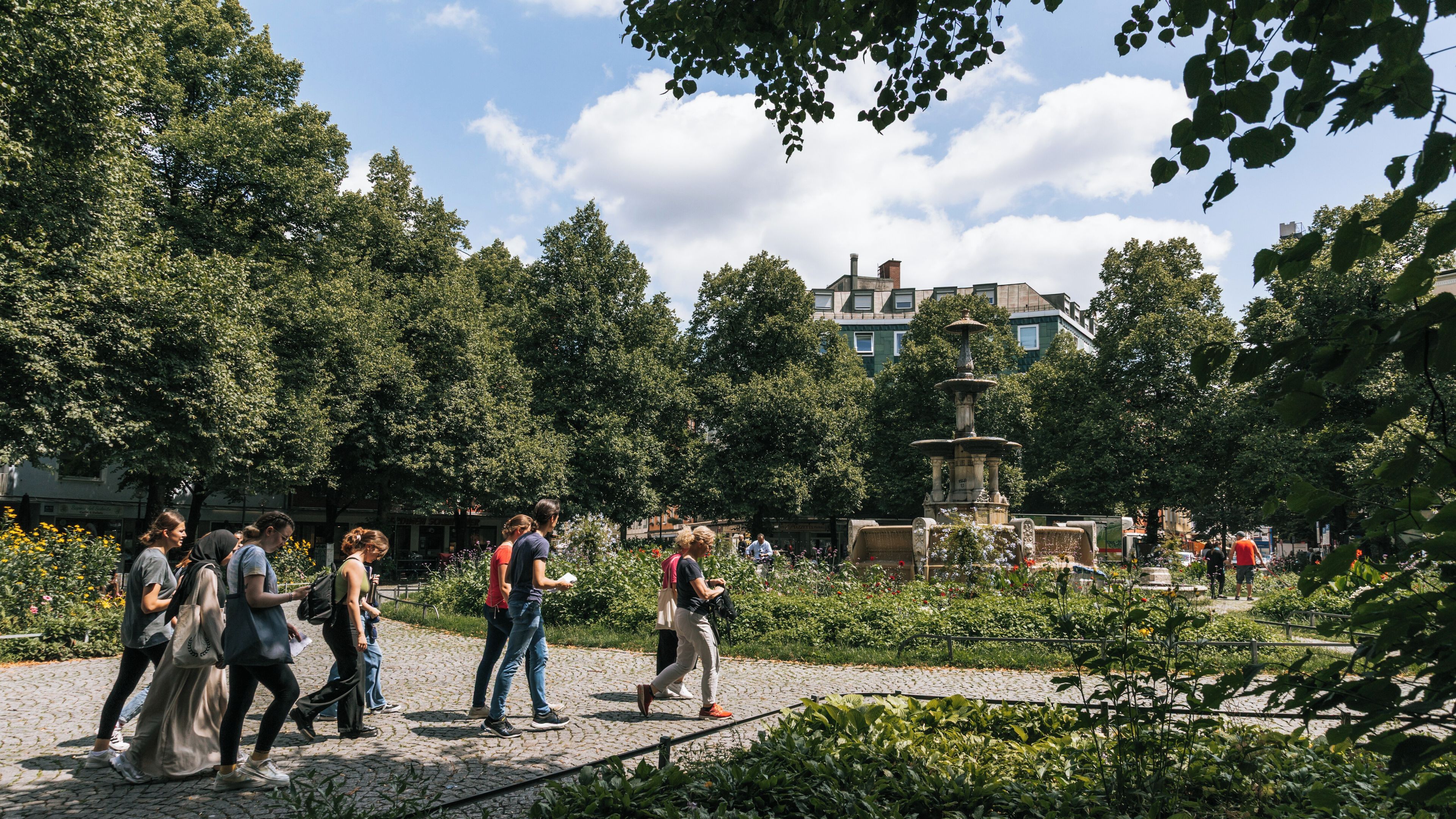 A park with a fountain, people walking, and trees.